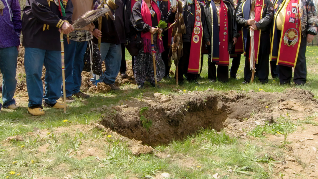 Aanikoobijigan image: Indigenous people gather around a grave, possibly for a traditional ceremony or memorial service.