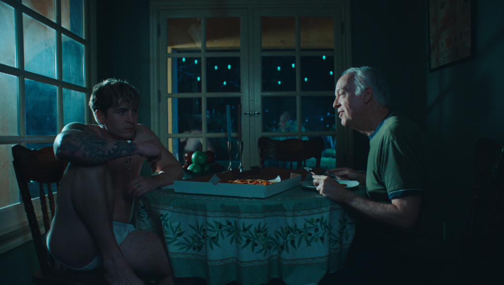 Still from Blue Film: Tattooed young man and older man share pizza in dimly lit scene, exploring themes of bravery and fear.