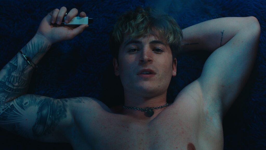 Blue Film still: Man with tattoos and necklace lies on blue surface, holding vape pen. Conversation with Elliot Tuttle & Kieron Moore.