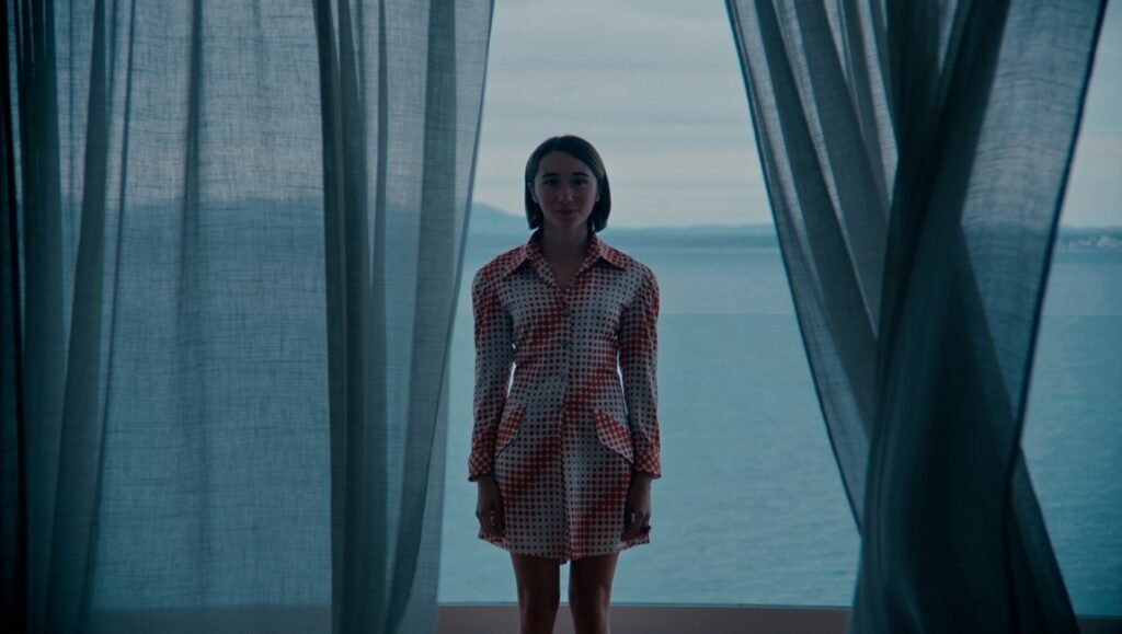 Brunette woman in a patterned dress stands between blue curtains framing a sea view.