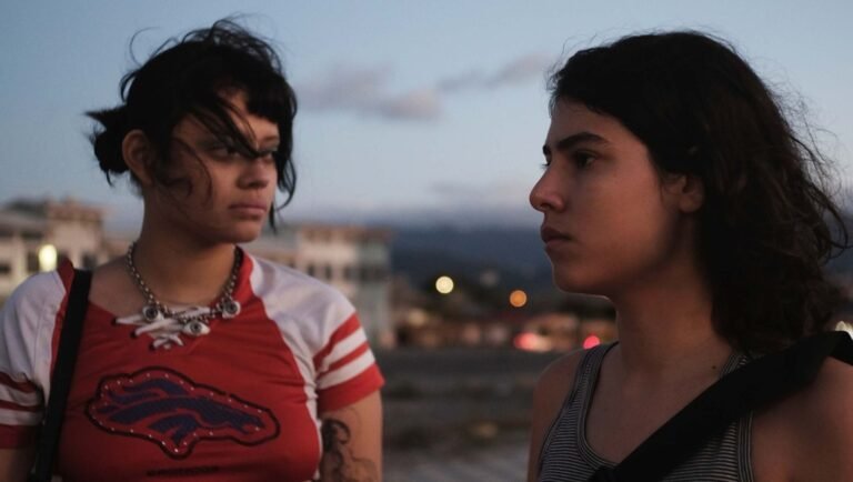 Two young women face each other outdoors at dusk, one in a red shirt with a Buffalo Bills logo and an eye-shaped necklace.