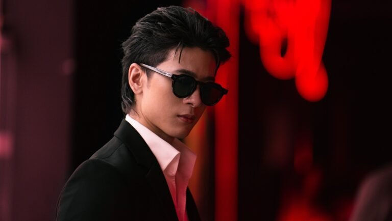 Man in a black suit and sunglasses poses against a dark backdrop with blurry red lighting.