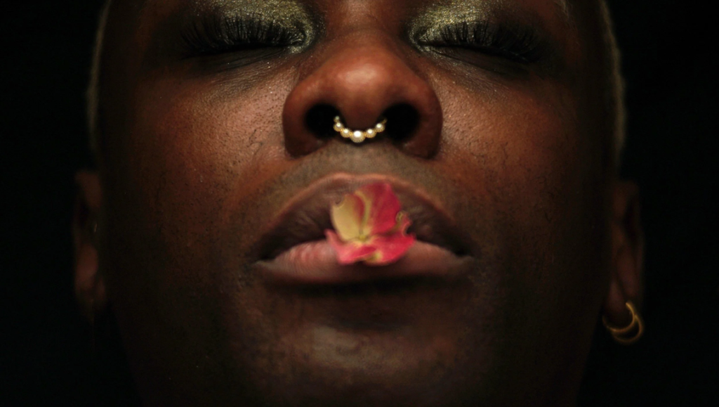 Close-up of person with eyes closed, flower in mouth, nose ring. "JoyBoy" stills for Kevin B. Lee's 'Afterlives' film review.