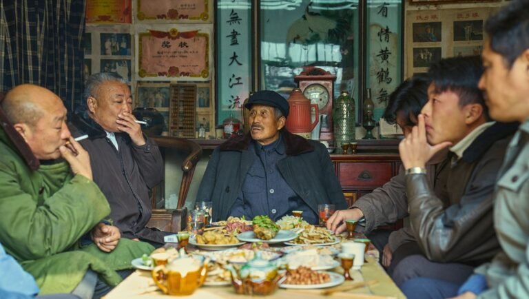 Living the Land film review: Group of men at a table laden with food and teapots, smoking cigarettes in an interior setting.
