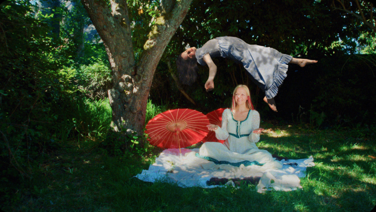 Woman levitates over a woman sitting on a blanket near two red parasols in a grassy area.