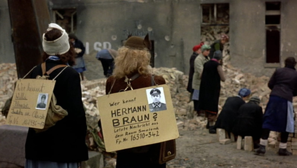 Two women stand among rubble, wearing signs with photos and text, including "Hermann Braun" and "Wer kennt.".