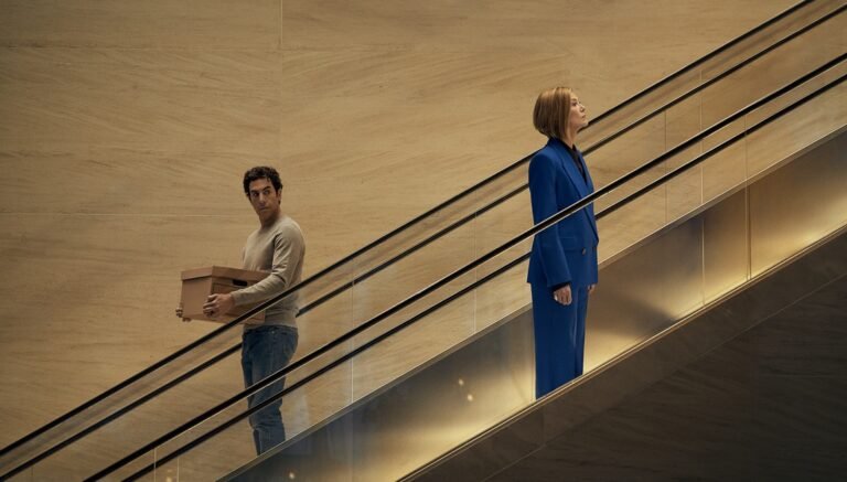 On an escalator, a man carries a box while a woman in a blue suit stands looking upward.
