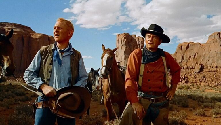 Two men stand with horses in a desert landscape, one in a black hat and red shirt, the other in a vest and blue neckerchief.