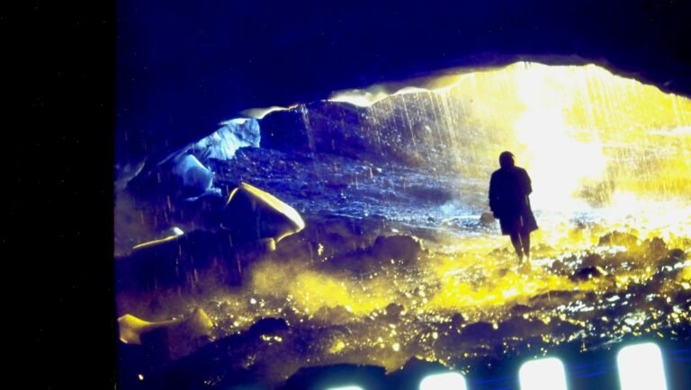 Person in dark coat stands in cave opening with bright light and water.