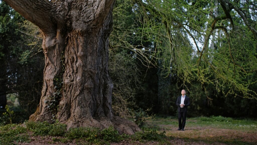 Tony Leung Chiu-wai in Silent Friend film, standing near a large tree, promoting Ildikó Enyedi's conversation.