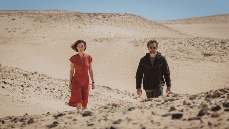 Woman in red dress and man in black jacket walk across sandy desert landscape.