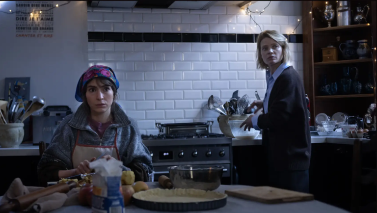 Two women prepare food in a kitchen with white subway tile and string lights.