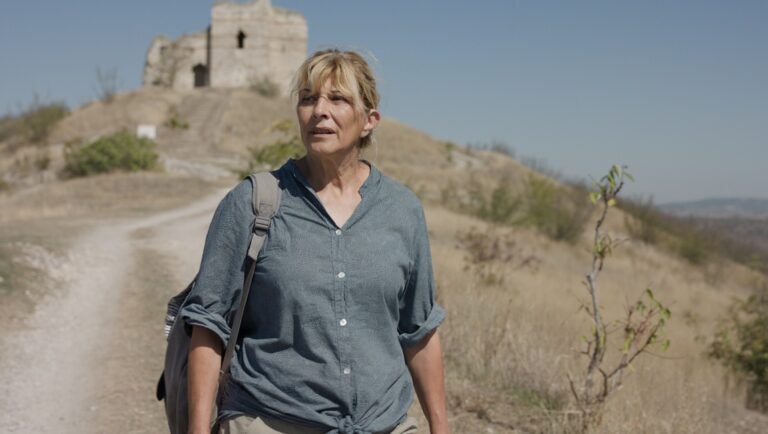 Woman with backpack hikes a dry grassy hillside toward a stone fortress ruin.