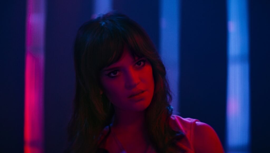 Woman with dark hair looks pensive under red and blue neon lighting.