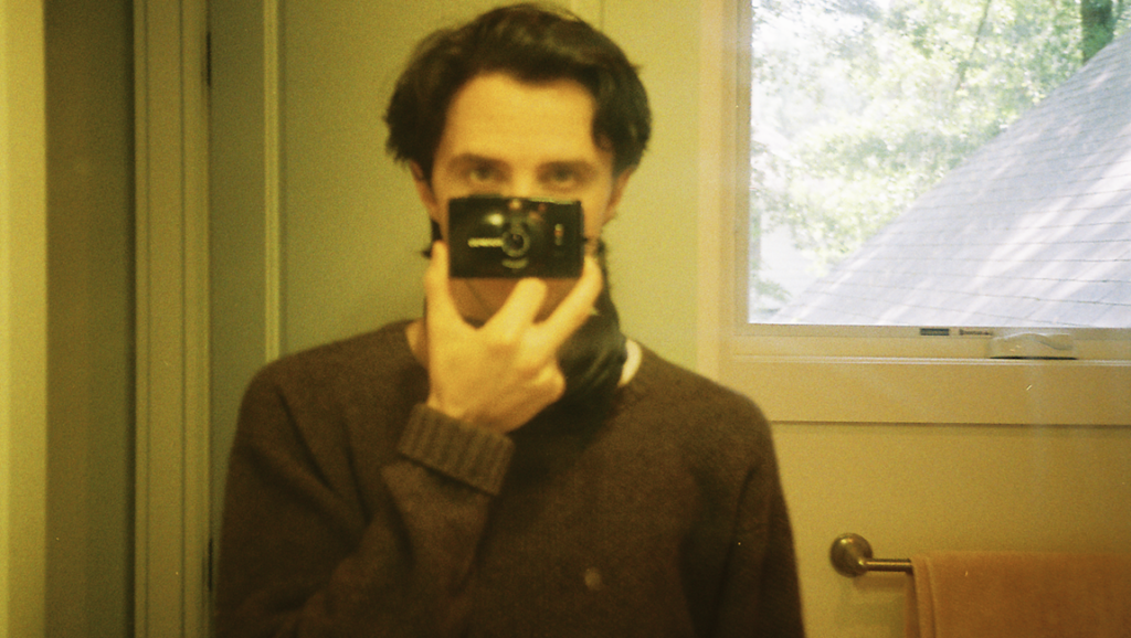 Josh Heaps, a photographer, takes a selfie in a bathroom, holding a camera to his face with a window in the background.