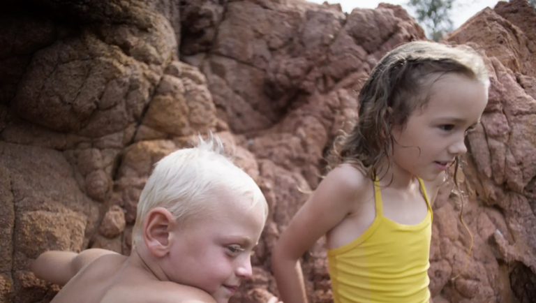 Two children stand near red rock formations; one wears a yellow swimsuit.