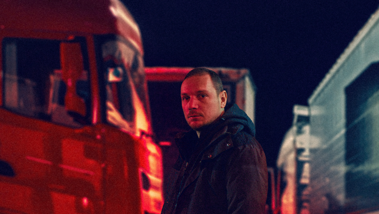 Man stands near a red truck at night under dark lighting.
