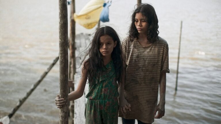 Two young girls stand on a wooden dock near the water, one in a floral romper and the other in a striped shirt.