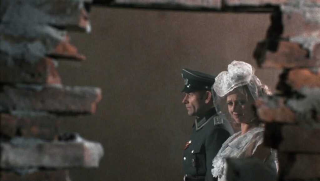 Woman in wedding dress and man in military uniform stand behind a broken brick wall.