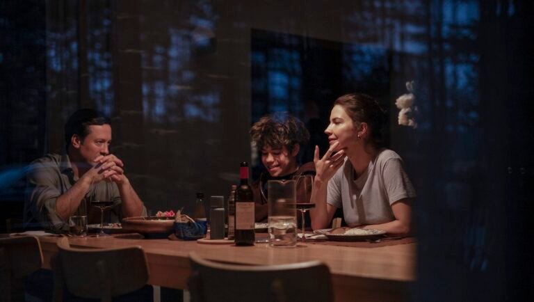 Family of three sits at a wooden table with wine and food, illuminated by dim interior lighting.