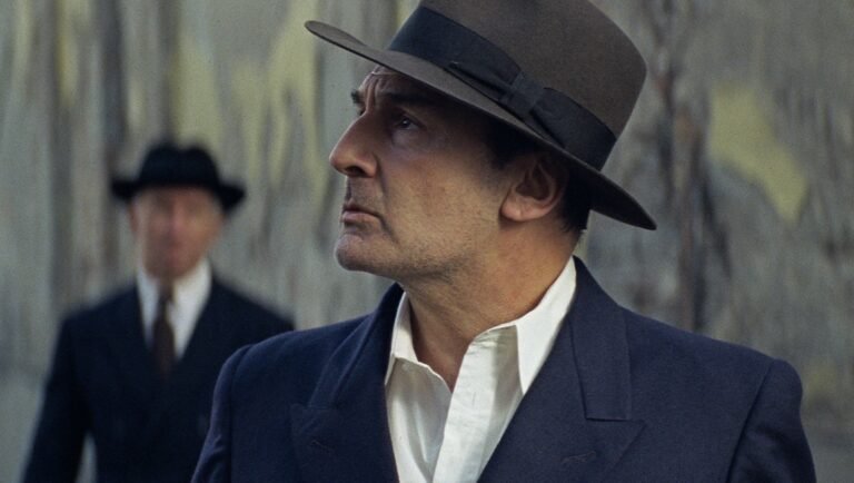 Profile of a man in a fedora and suit, gazing upwards, with another suited figure blurred in the background.