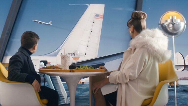 People sit at a table near an airport window with a jet on the tarmac, and a plane in the sky.