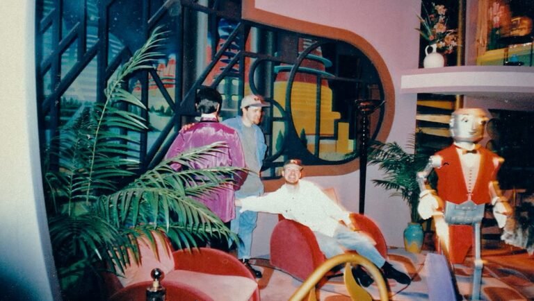 Three men pose in a room with a robot butler, stylized city backdrop, and various palm plants.
