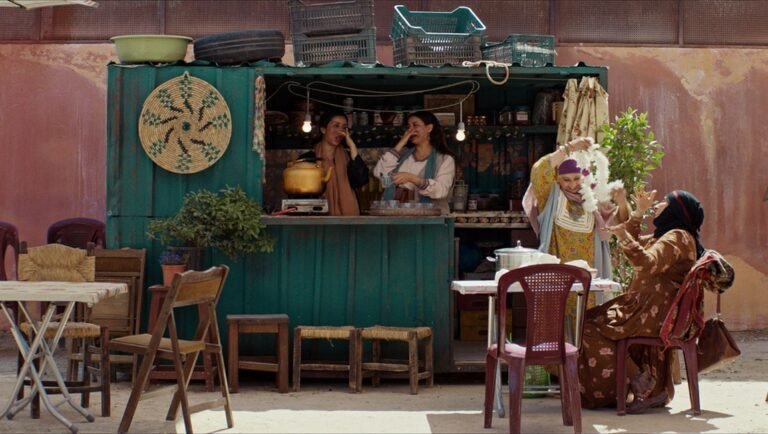 Women at a roadside stand with food and hanging lights prepare a flower garland for a seated woman.