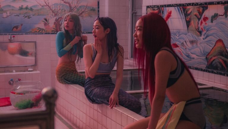 Three women wearing mermaid tails sit in a tiled bathroom with a fishbowl and a mural of koi fish.