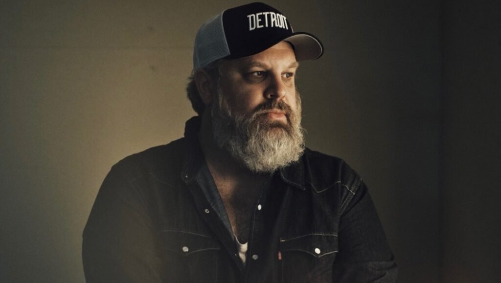 Adam Carter Rehmeier with a full beard wearing a black Detroit trucker hat and a dark denim shirt.