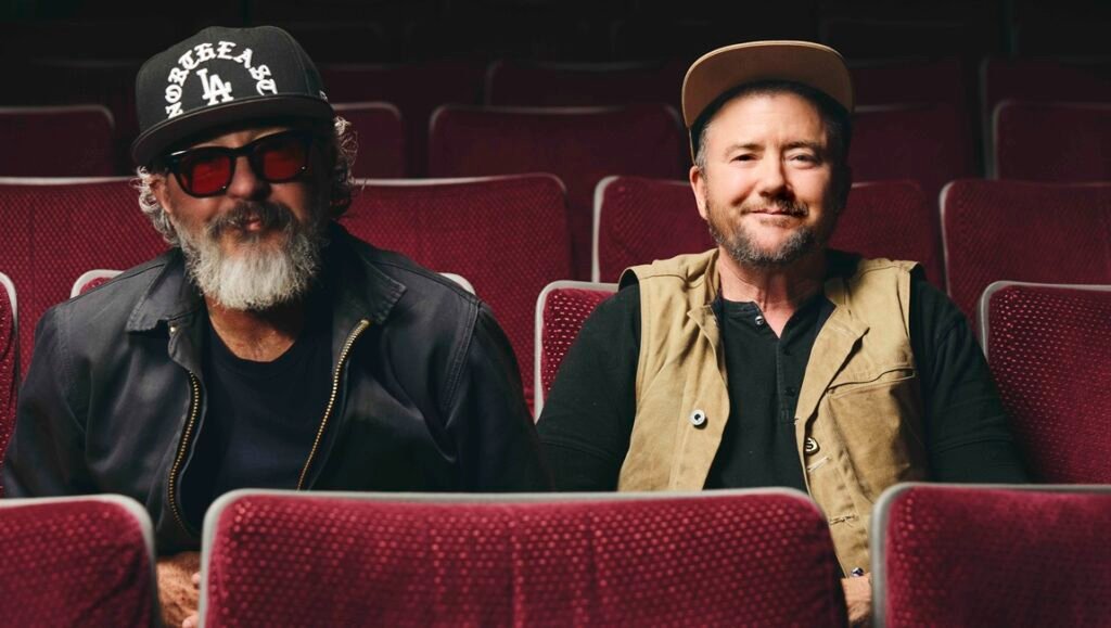 Harry Dodge and Silas Howard sit in red theater seats, both wearing hats and casual jackets.