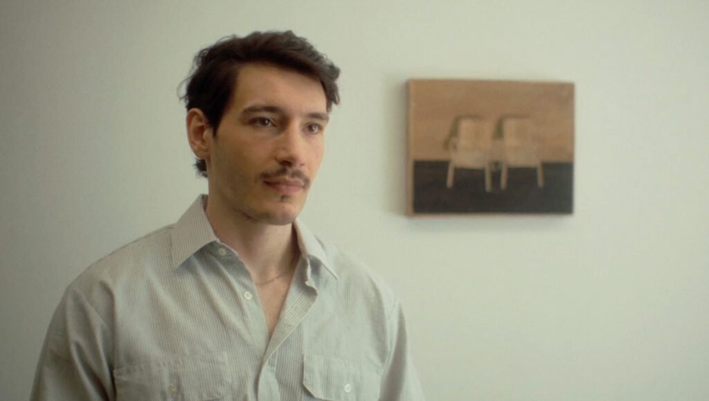 Young man with short dark hair and a mustache wearing a light-colored button-down shirt standing before a white wall.