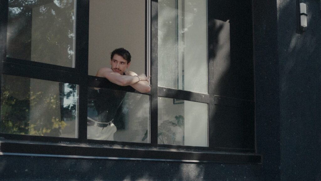 Man with a mustache wearing a black tank top leaning on an open window frame while looking outside.
