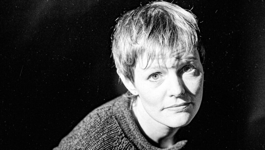 Close-up portrait of a woman with short hair wearing a knitted sweater against a dark background.