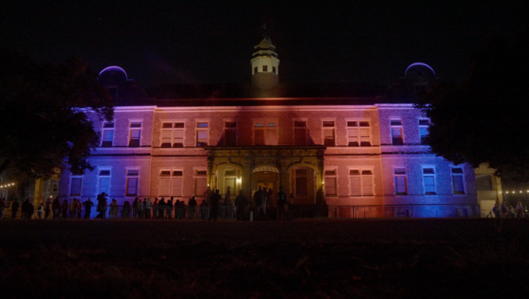 Crowd gathers before a grand brick building illuminated by dramatic red and blue architectural lighting at night.