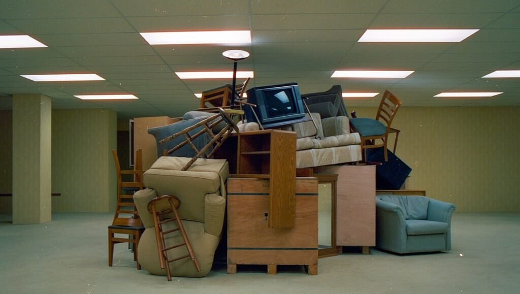 Large chaotic pile of mismatched furniture, chairs, and an old television in a dimly lit, yellow-walled office space.