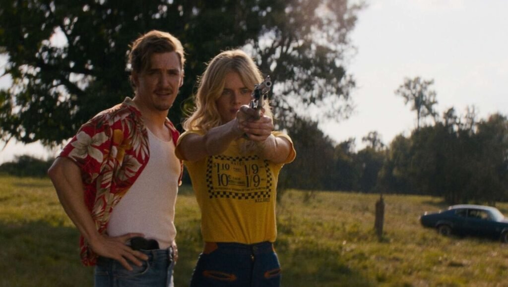 Blonde woman in a yellow shirt aims a handgun while a man in a red floral shirt stands beside her in a rural field.