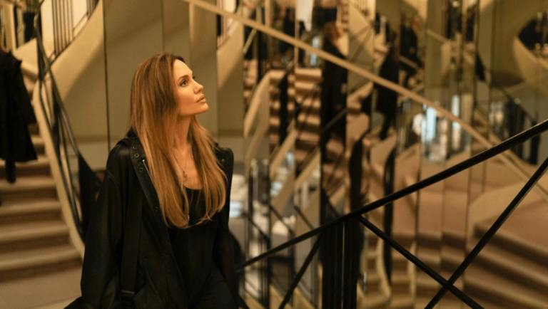 Angelina Jolie with long blonde hair walking up a curved staircase in a room with mirrored walls.