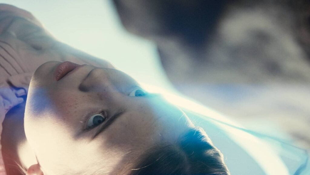 A young child with freckles looks upward with wide blue eyes, illuminated by a bright, ethereal blue light.