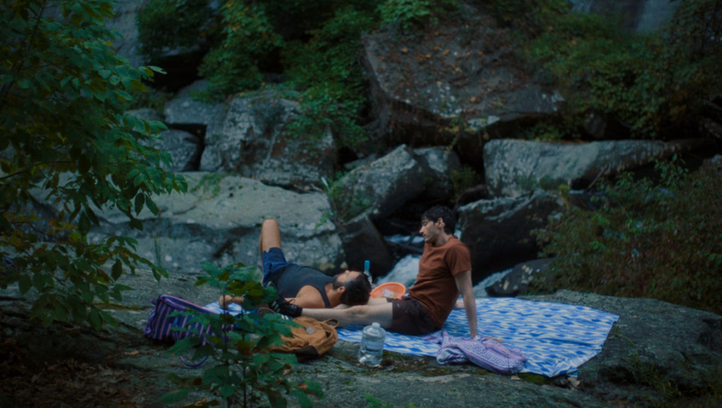 Two men relaxing on a blue patterned blanket atop a rocky ledge near a forested stream.