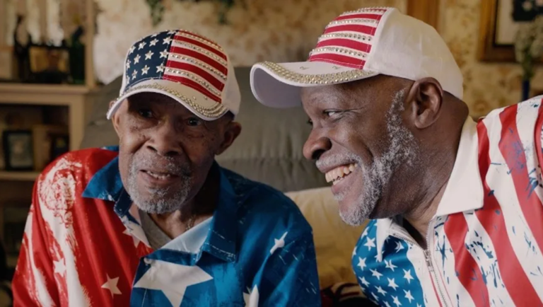 Two smiling Black men wearing matching American flag-patterned shirts and rhinestone-embellished caps.