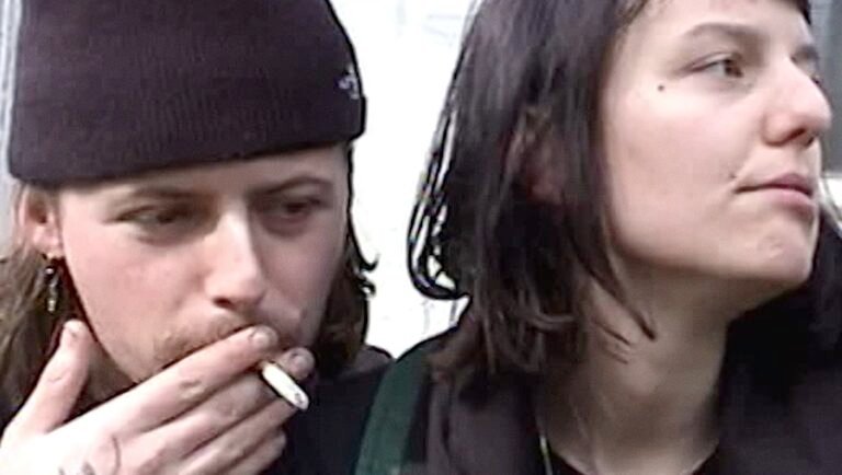 Young man in a black beanie smokes a cigarette beside a dark-haired woman looking away.