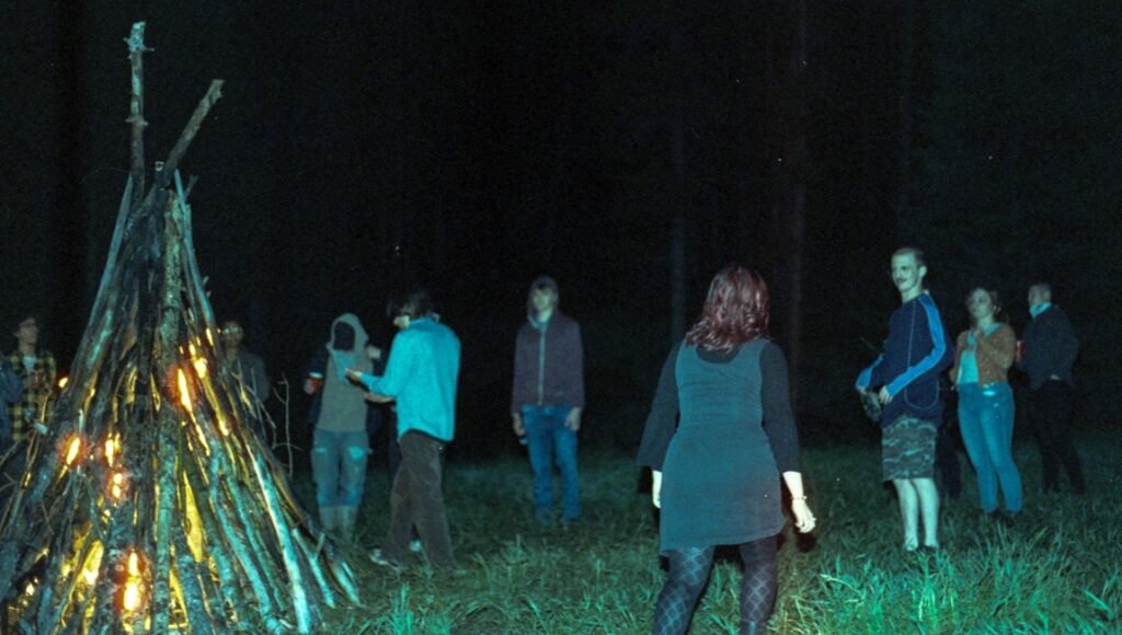 Group of people stands around a tall, glowing campfire in a dark, wooded clearing at night.