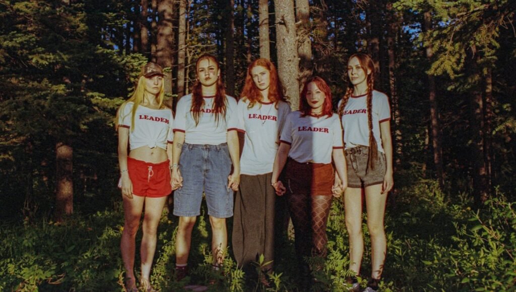 Five young women holding hands in a line, wearing white LEADER t-shirts, stand amidst pine trees in a forest at sunset.