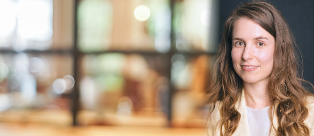 Sophia Takal smiles gently while looking at the camera against a blurred indoor backdrop.