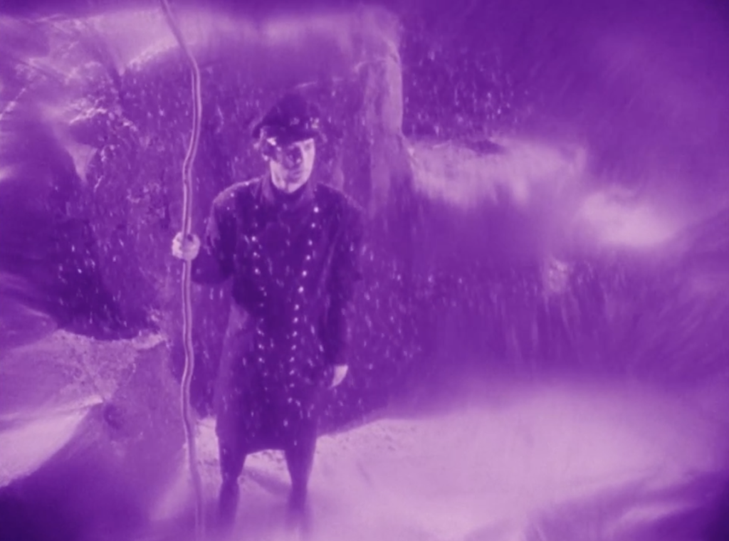 Uniformed individual holding a long staff stands in a mist-filled cavern illuminated by saturated violet light.