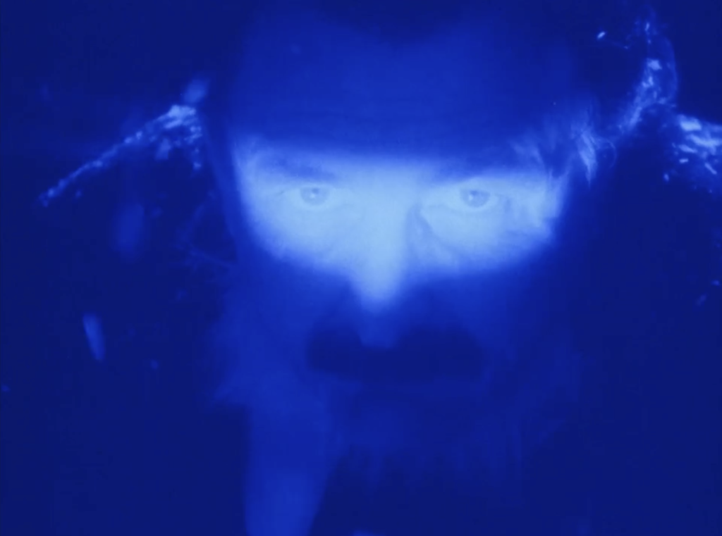Close-up portrait of a man with an intense gaze illuminated by vibrant blue light against a dark background.