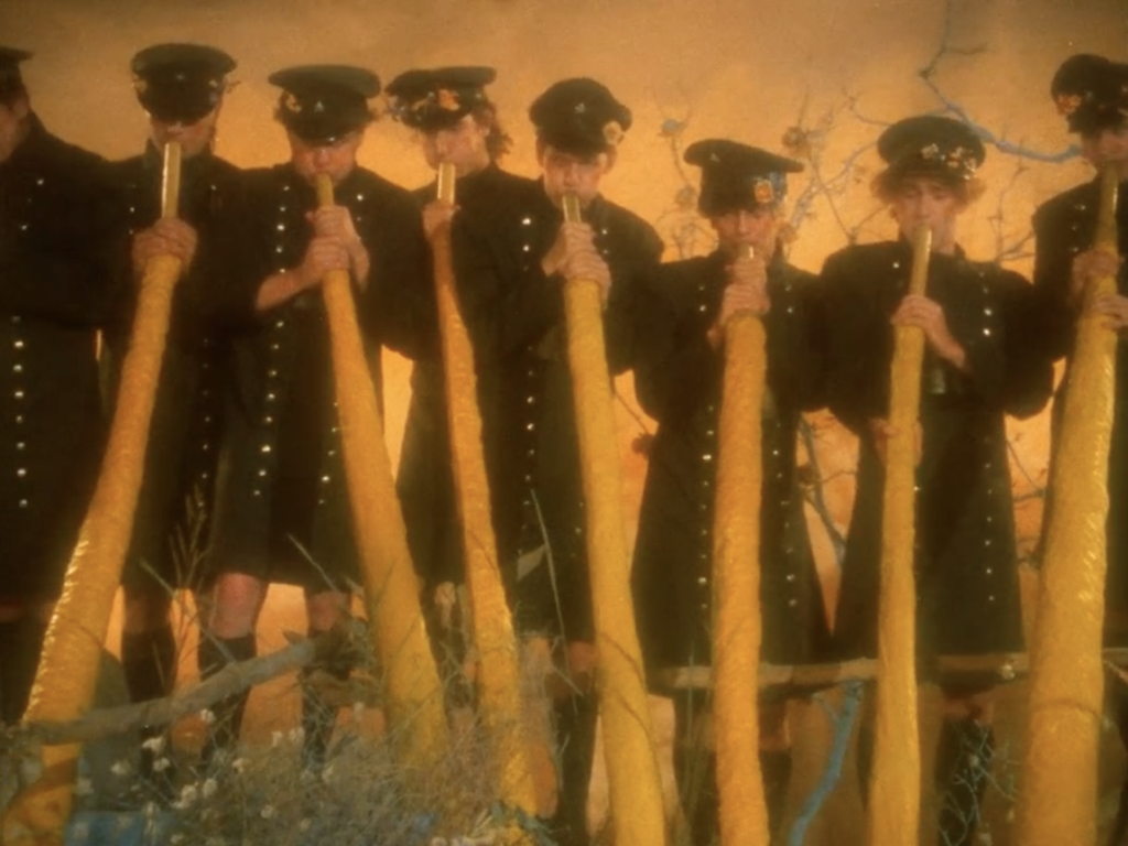 Group of men in dark military-style uniforms and caps playing long, vertical yellow alphorns against a golden background.