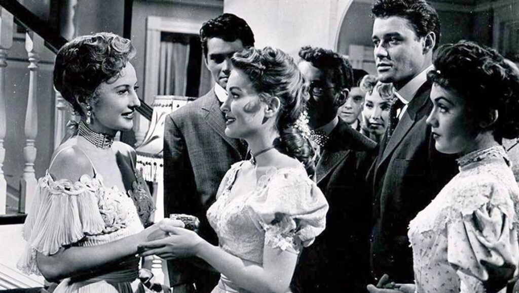 Barbara Stanwyck in a ruffled gown greets a young woman at a formal gathering surrounded by men in suits.