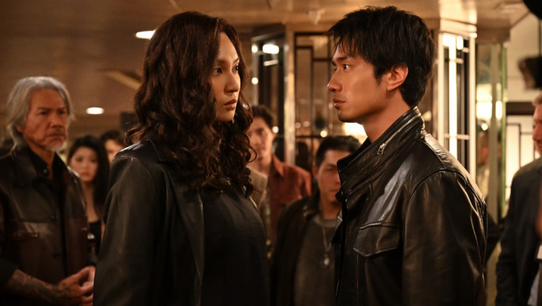 A man and woman in leather jackets stand face-to-face in a tense confrontation surrounded by a crowd.